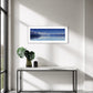 A framed panoramic landscape print of Aoraki Mt Cook reflected in a calm blue waters of Lake Pukaki hangs on a white wall above a marble console table with a green plant and a small black speaker in a modern, sunlit room.