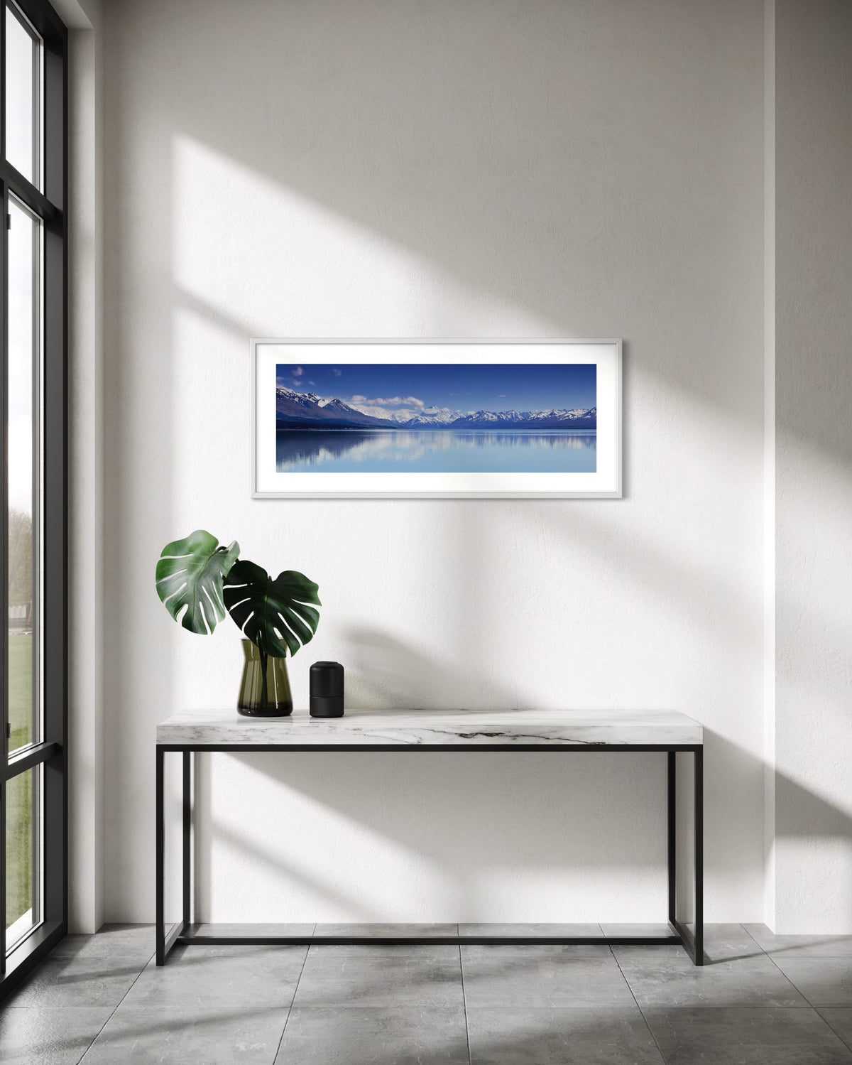 A framed panoramic landscape print of Aoraki Mt Cook reflected in a calm blue waters of Lake Pukaki hangs on a white wall above a marble console table with a green plant and a small black speaker in a modern, sunlit room.