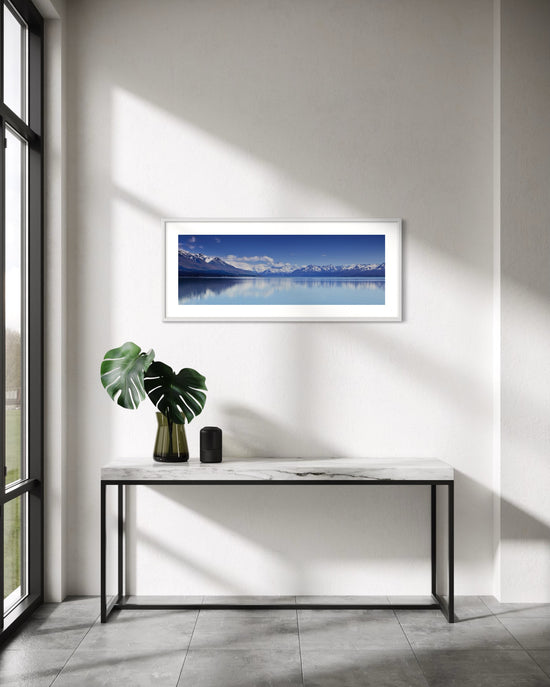A framed panoramic landscape print of Aoraki Mt Cook reflected in a calm blue waters of Lake Pukaki hangs on a white wall above a marble console table with a green plant and a small black speaker in a modern, sunlit room.