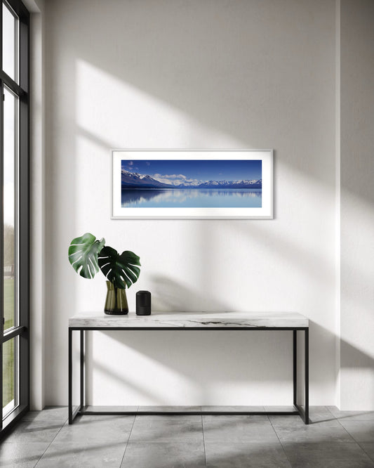 A framed panoramic landscape print of Aoraki Mt Cook reflected in a calm blue waters of Lake Pukaki hangs on a white wall above a marble console table with a green plant and a small black speaker in a modern, sunlit room.