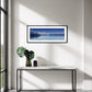 A framed horizontal landscape photograph of Aoraki Mt Cook  reflected in a calm blue waters of Lake Pukaki, displayed on a white wall above a modern console table with a green plant in a vase.