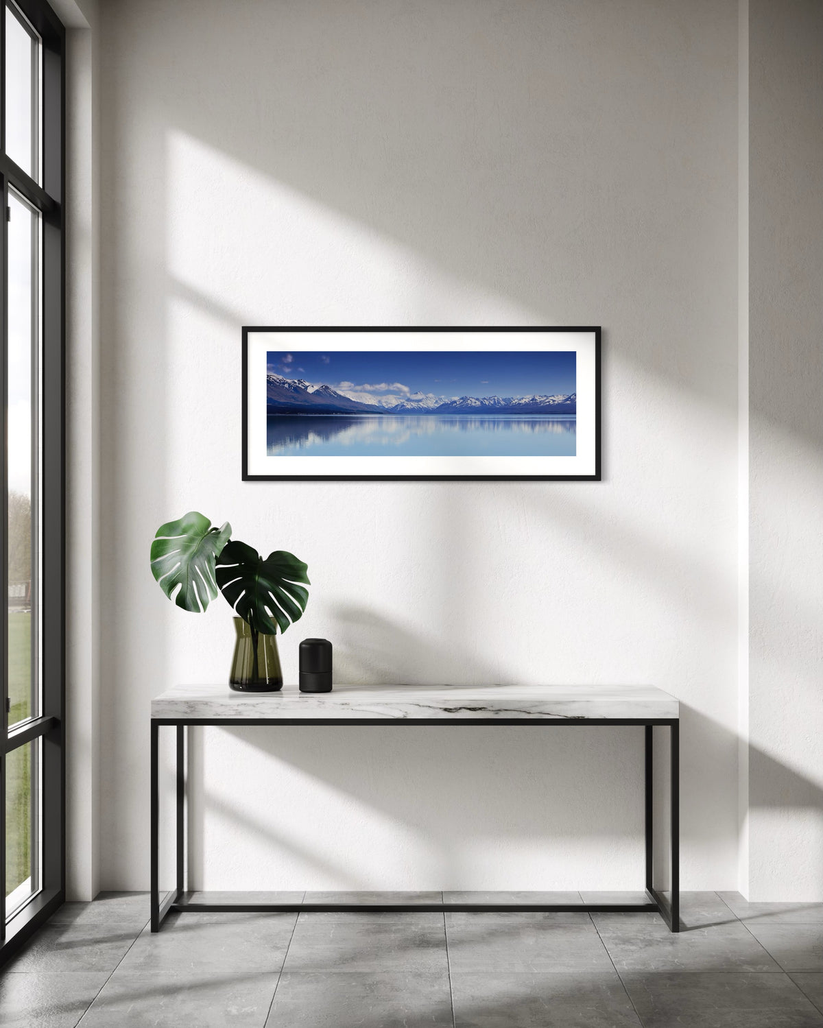 A framed horizontal landscape photograph of Aoraki Mt Cook  reflected in a calm blue waters of Lake Pukaki, displayed on a white wall above a modern console table with a green plant in a vase.
