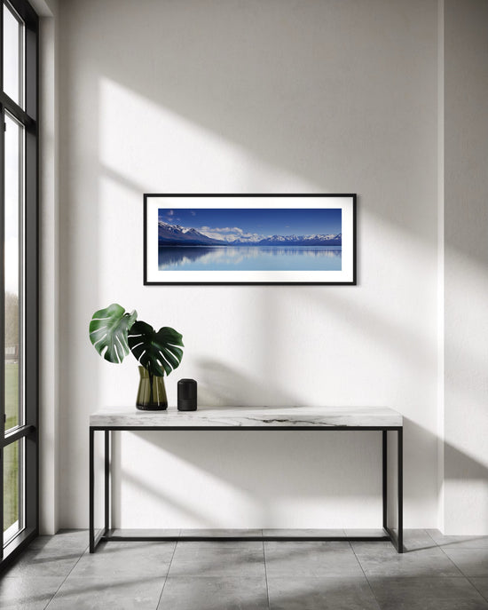 A framed horizontal landscape photograph of Aoraki Mt Cook  reflected in a calm blue waters of Lake Pukaki, displayed on a white wall above a modern console table with a green plant in a vase.